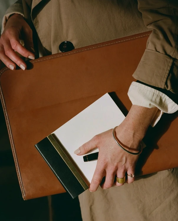 Hands in a camel coat sleeve holding a tan leather folio and notebook